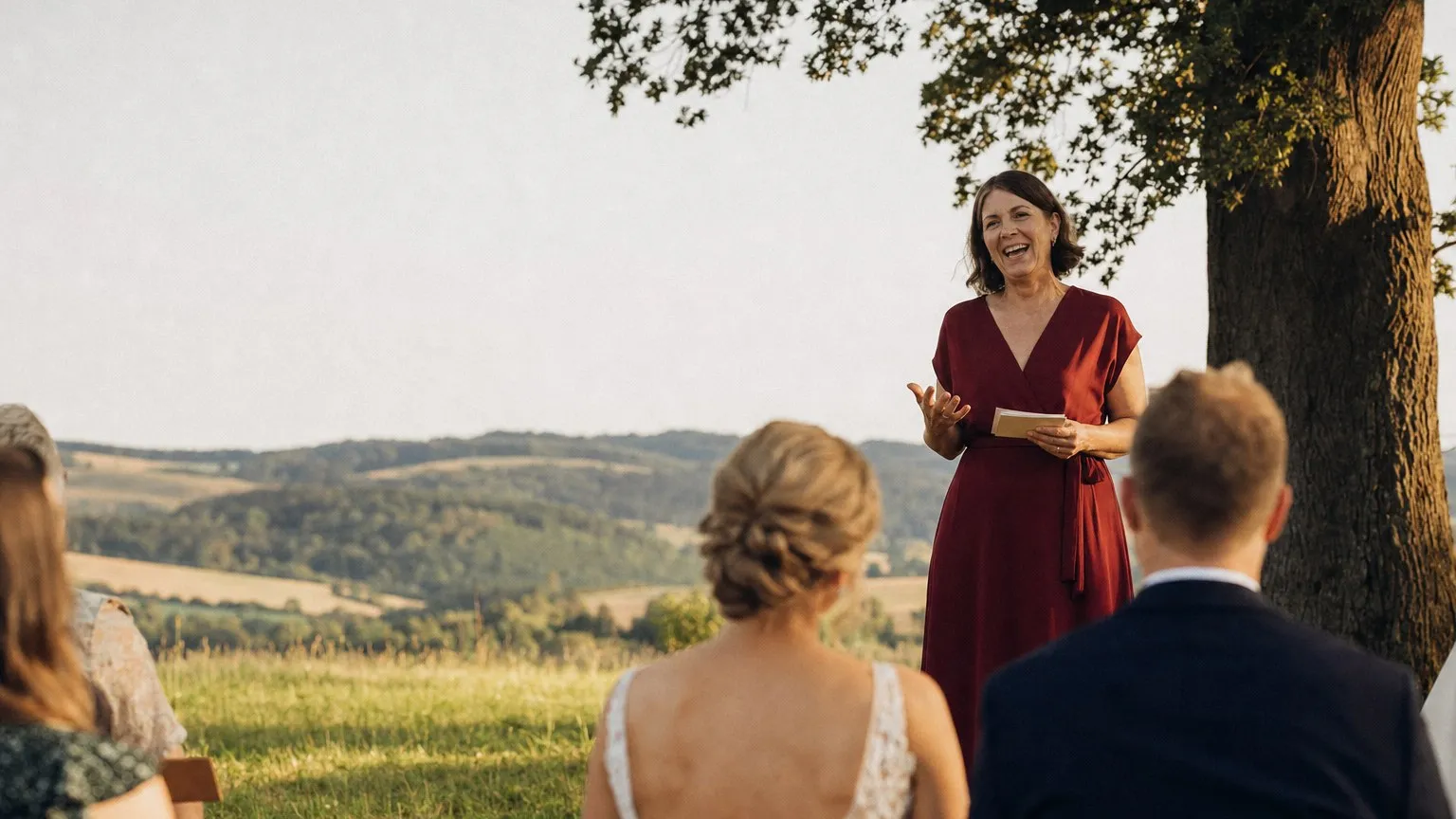 Eine Traurednerin im dunkelroten Kleid spricht lachend bei einer freien Trauung unter einer Eiche, das Brautpaar steht im Vordergrund.